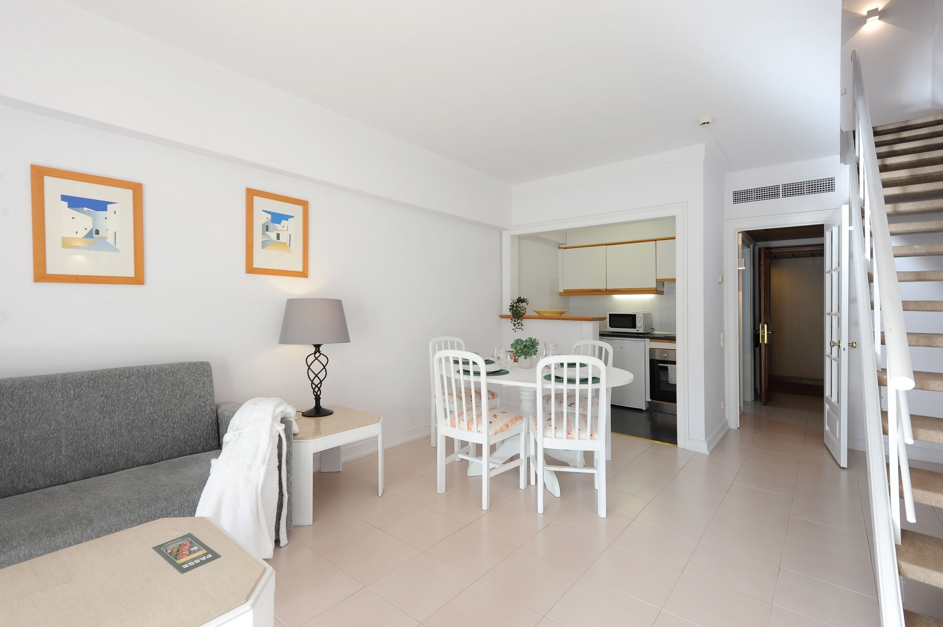 Uma sala de estar moderna com um sofá cinzento, uma pequena mesa com uma luminária e um espaço de refeições com cadeiras brancas e uma mesa. É visível uma cozinha ao fundo.