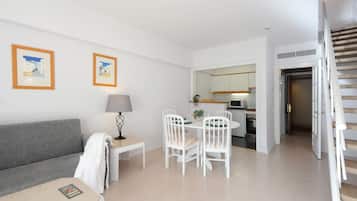 Uma sala de estar moderna com um sofá cinzento, uma pequena mesa com uma luminária e um espaço de refeições com cadeiras brancas e uma mesa. É visível uma cozinha ao fundo.