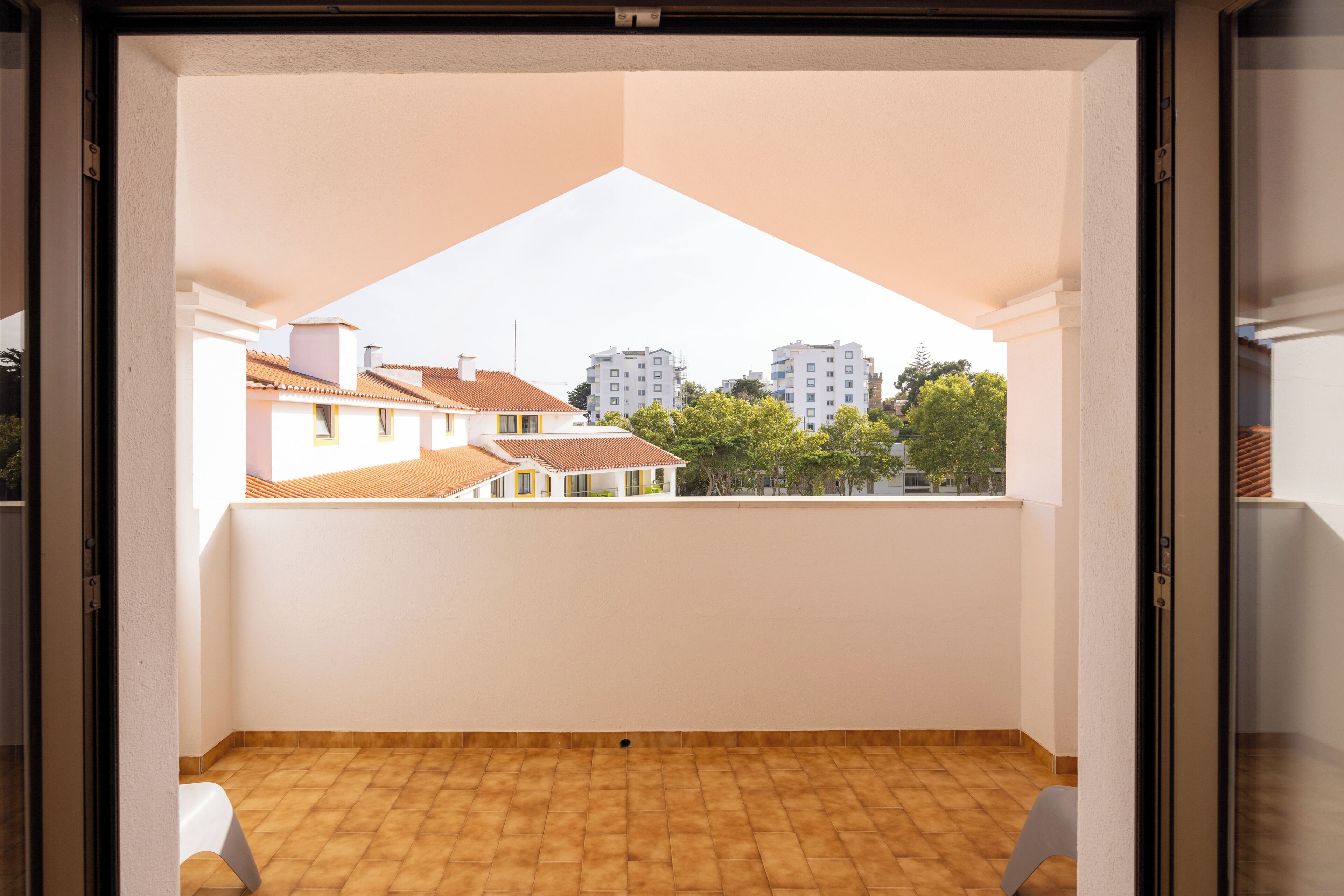 Un balcon doté d’un sol carrelé, de murs blancs et offrant une vue sur les bâtiments et les arbres environnants.