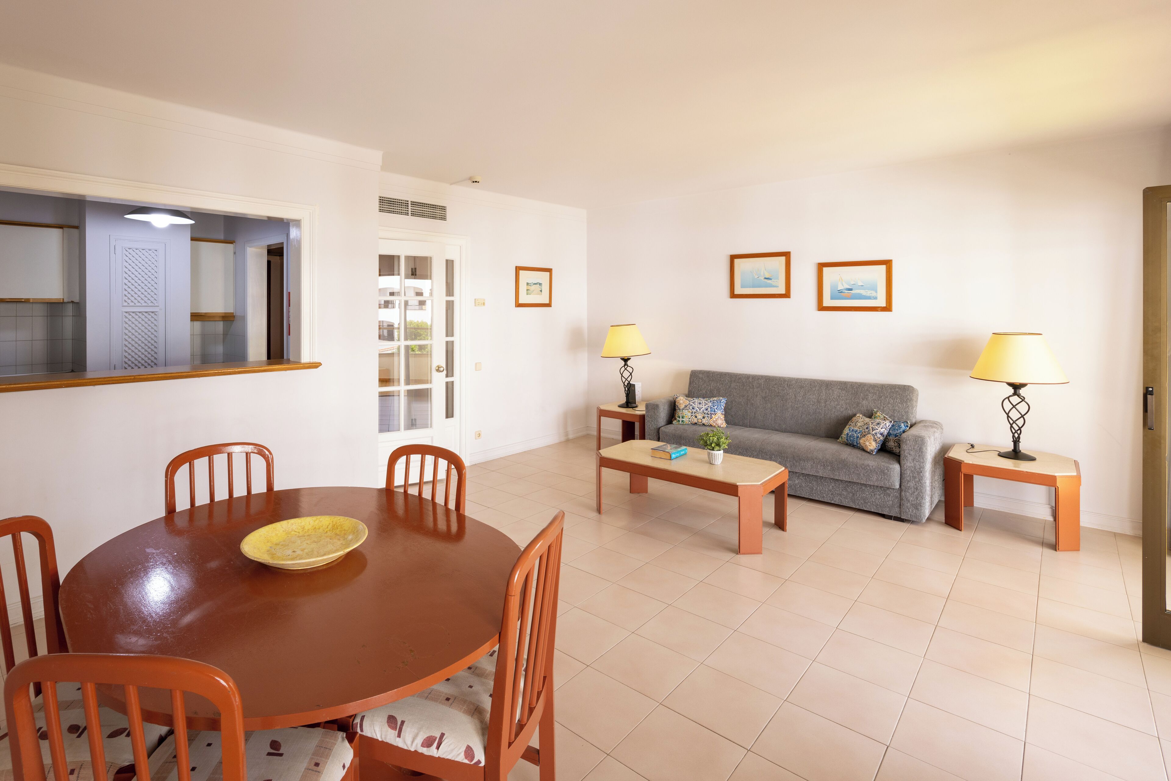 Uma sala de estar com um sofá cinzento, duas mesas laterais com luminárias, uma mesa de centro e um espaço de jantar com uma mesa redonda e cadeiras.