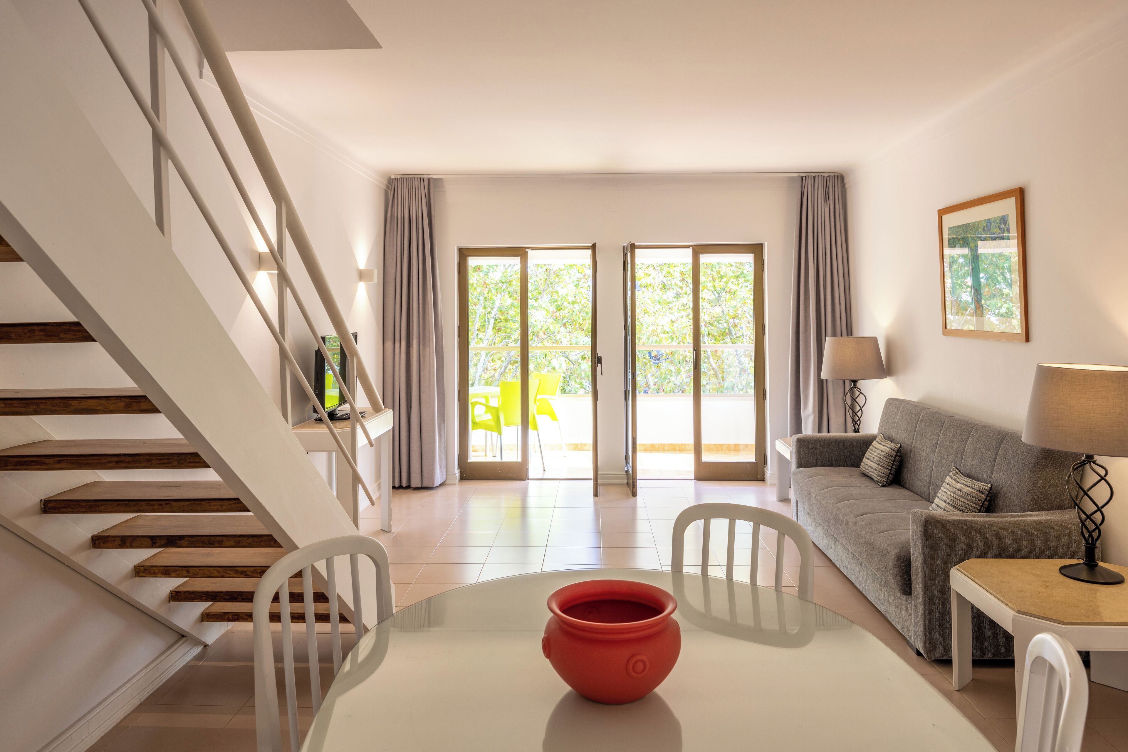 A modern living room with a staircase, a dining table with a red pot, a grey sofa, and a lamp.