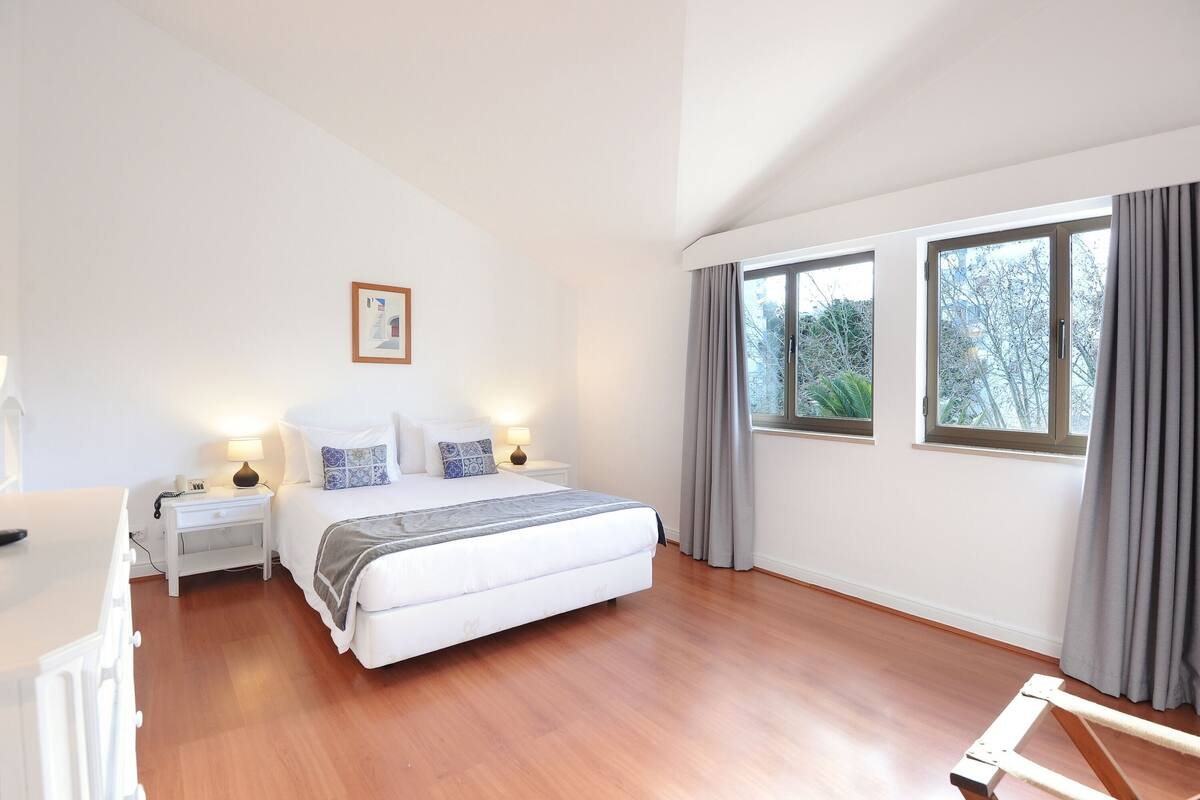 A bedroom with a bed, bedside tables, a nightstand, and two windows with curtains.