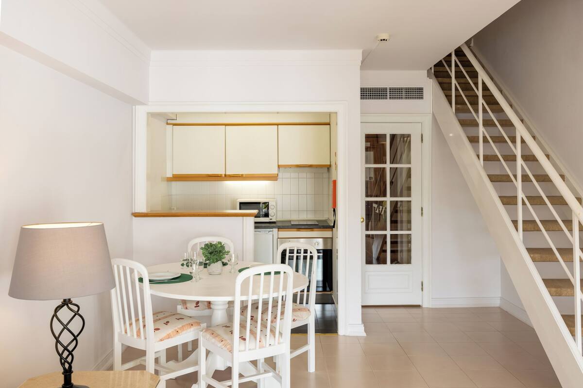 A dining area with a round table, white chairs, and a lamp. There is a kitchen visible through an open door.