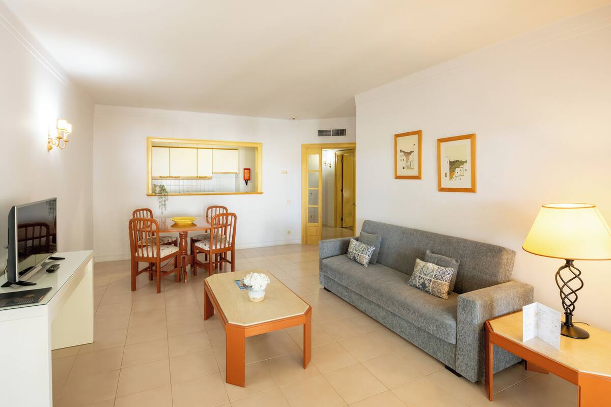 A living room with a sofa, coffee table, and dining table with chairs. There is a TV on a stand, a lamp, and framed pictures on the wall.