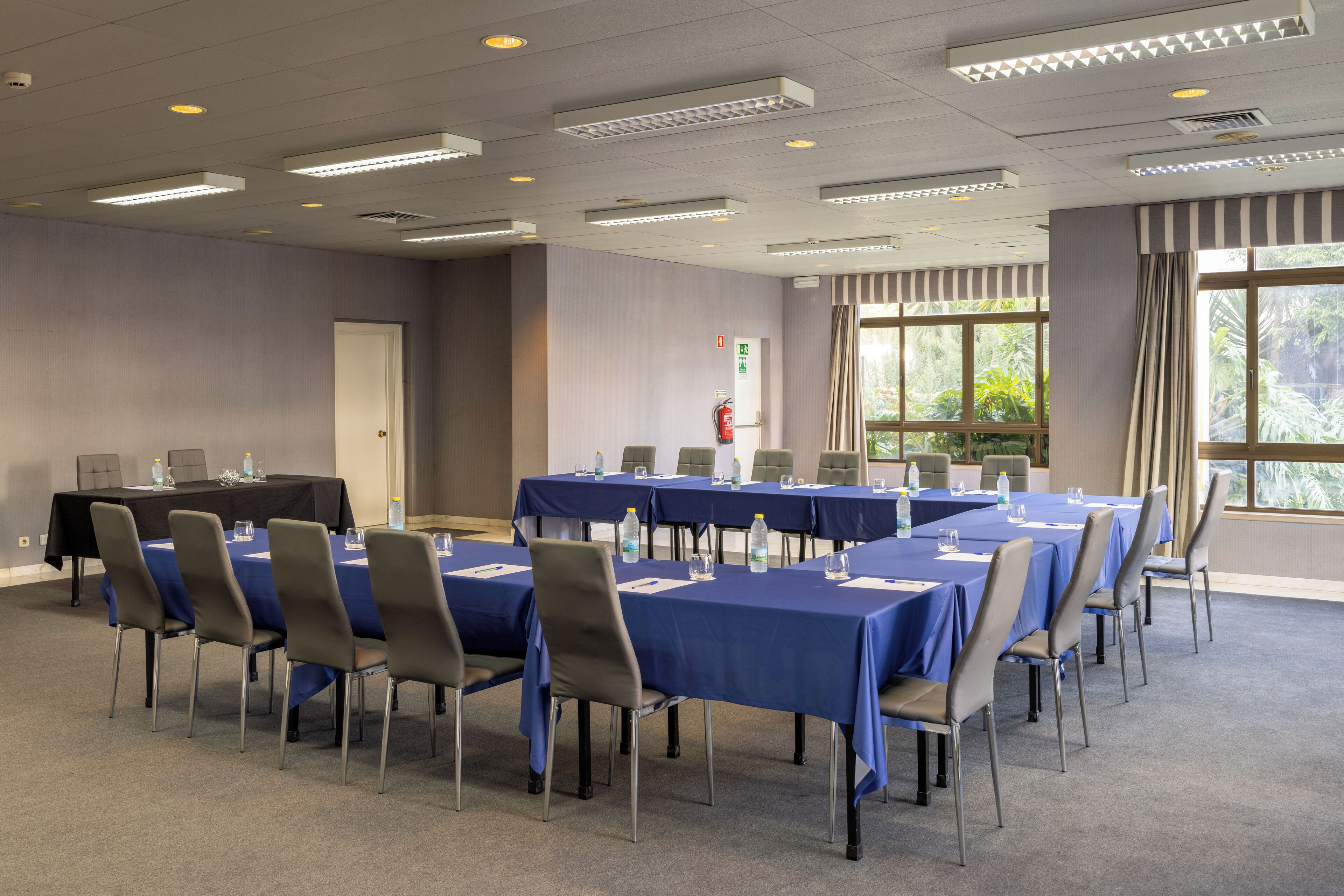 Sala de conferência com várias mesas e cadeiras, equipada com garrafas de água e cadernos.