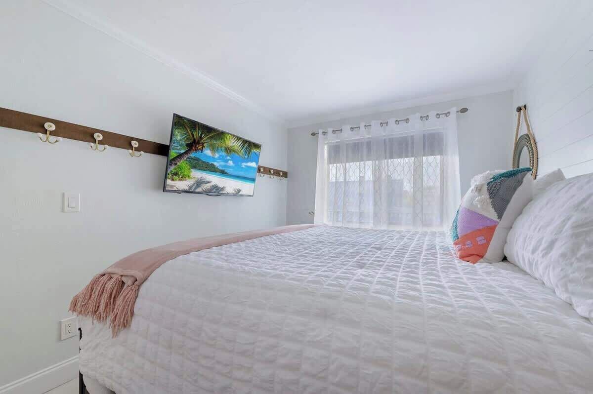 A bedroom with a bed, a painting of a beach scene, a window with sheer curtains, and a wall-mounted coat rack.