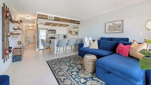 A modern living room with a blue sofa, a patterned rug, and a kitchen in the background. - Modern Condo With Pool Steps From Vanderbilt Beach (Naples)