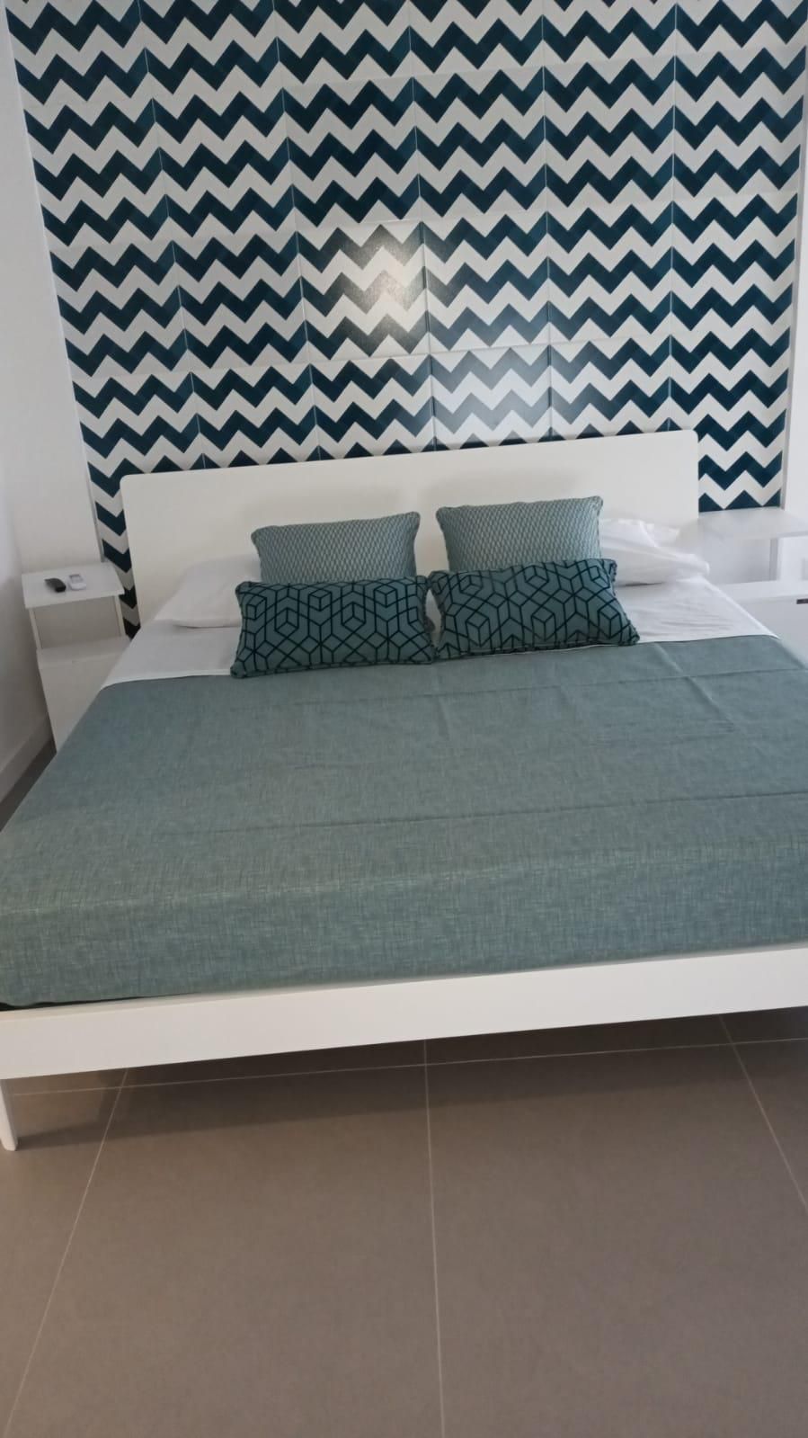 A bed with a teal bedspread and two patterned pillows, a white headboard, and a wall with a zigzag pattern.
