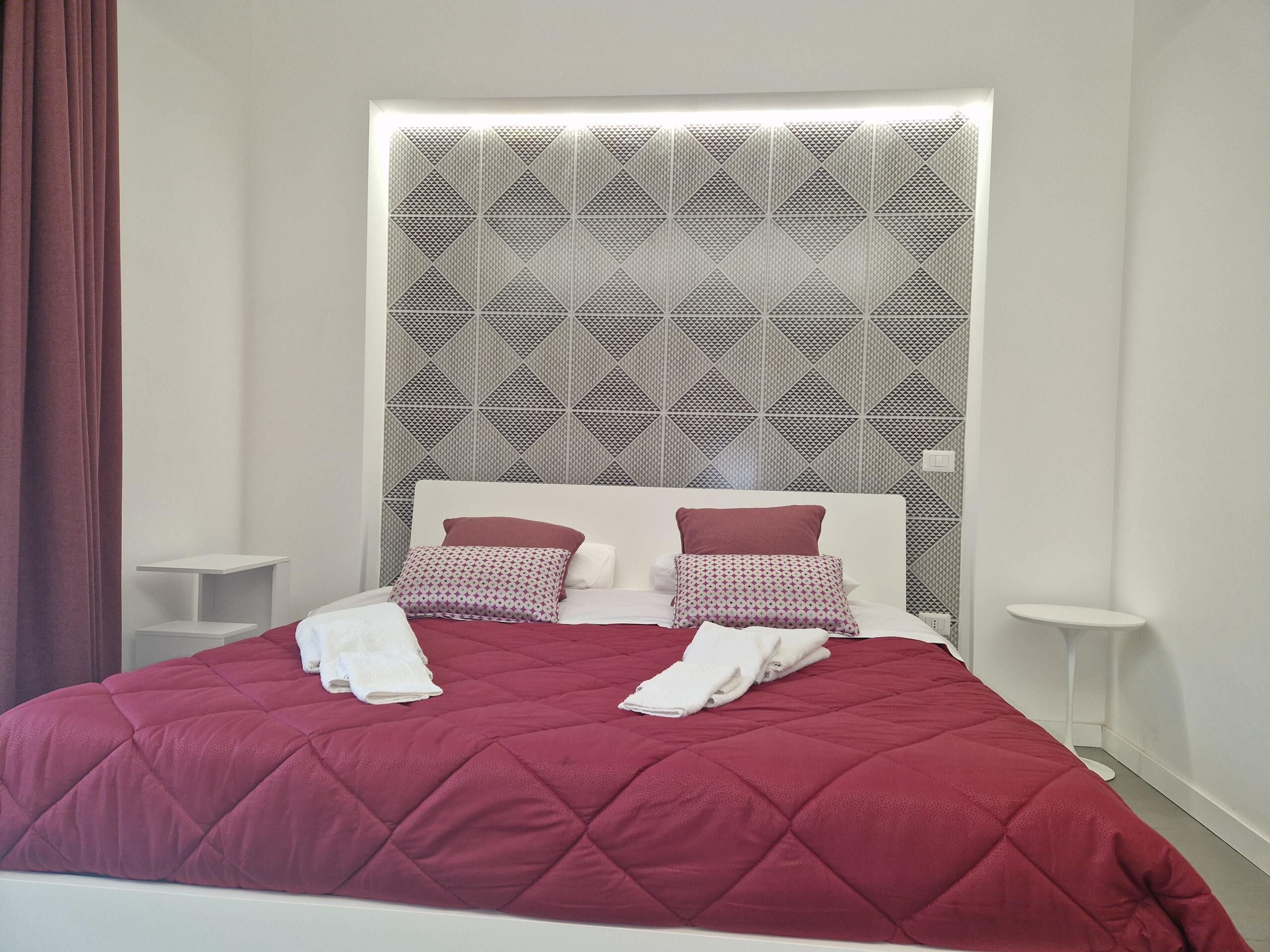Una cama bien tendida con colcha roja, dos almohadas y cabecera blanca.