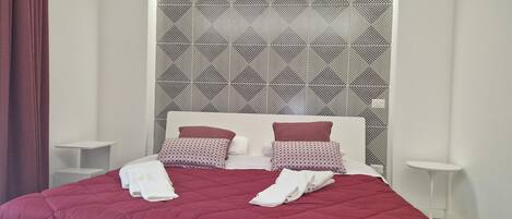 Una cama bien tendida con colcha roja, dos almohadas y cabecera blanca.