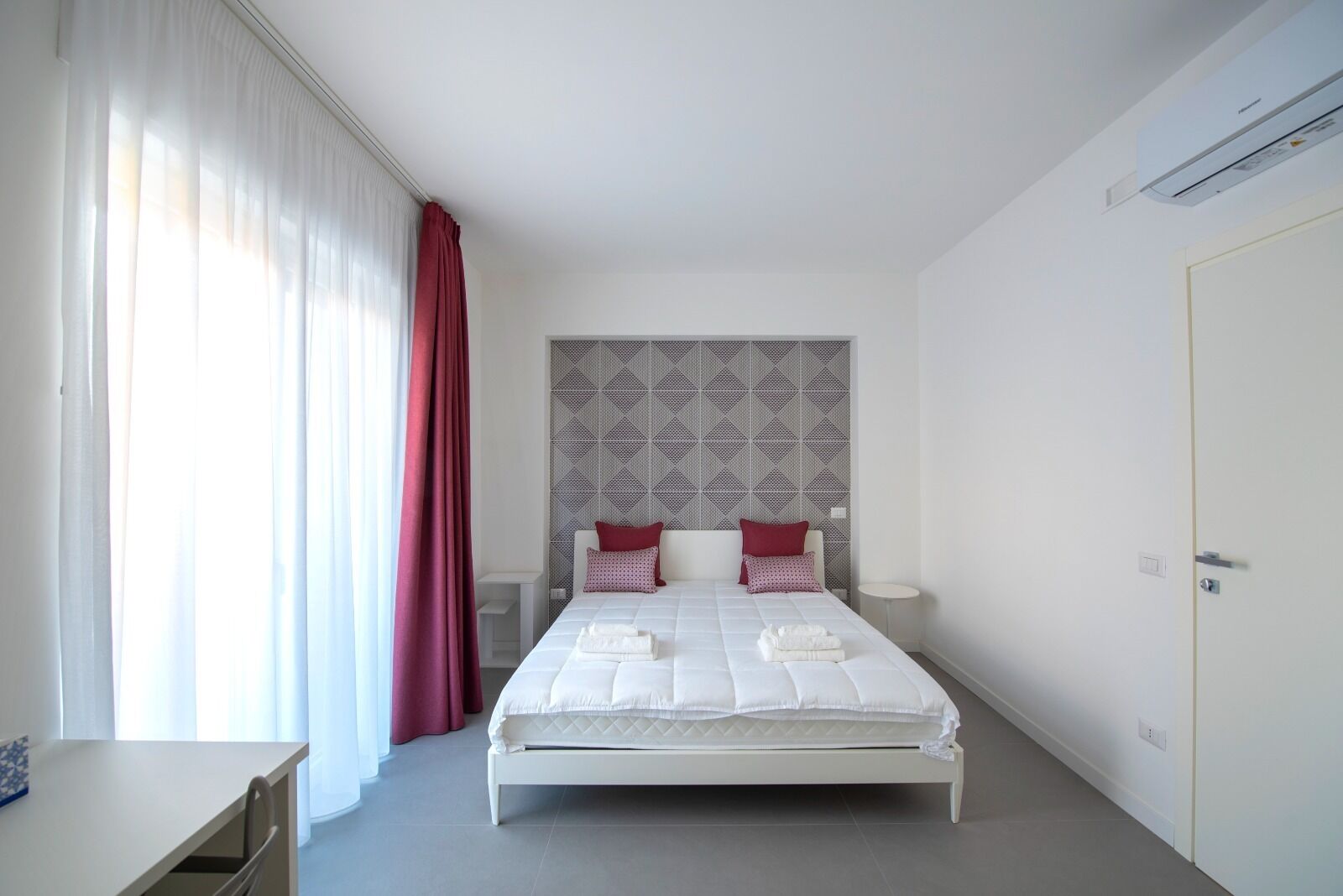 A modern bedroom with a white bed, red curtains, a geometric-patterned headboard, and a small bedside table.