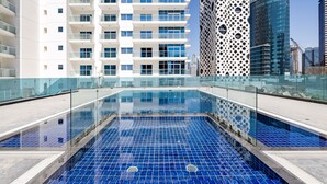 Una piscina en la azotea con barandillas de vidrio transparente, rodeada de edificios modernos con balcones y una fachada perforada distintiva.