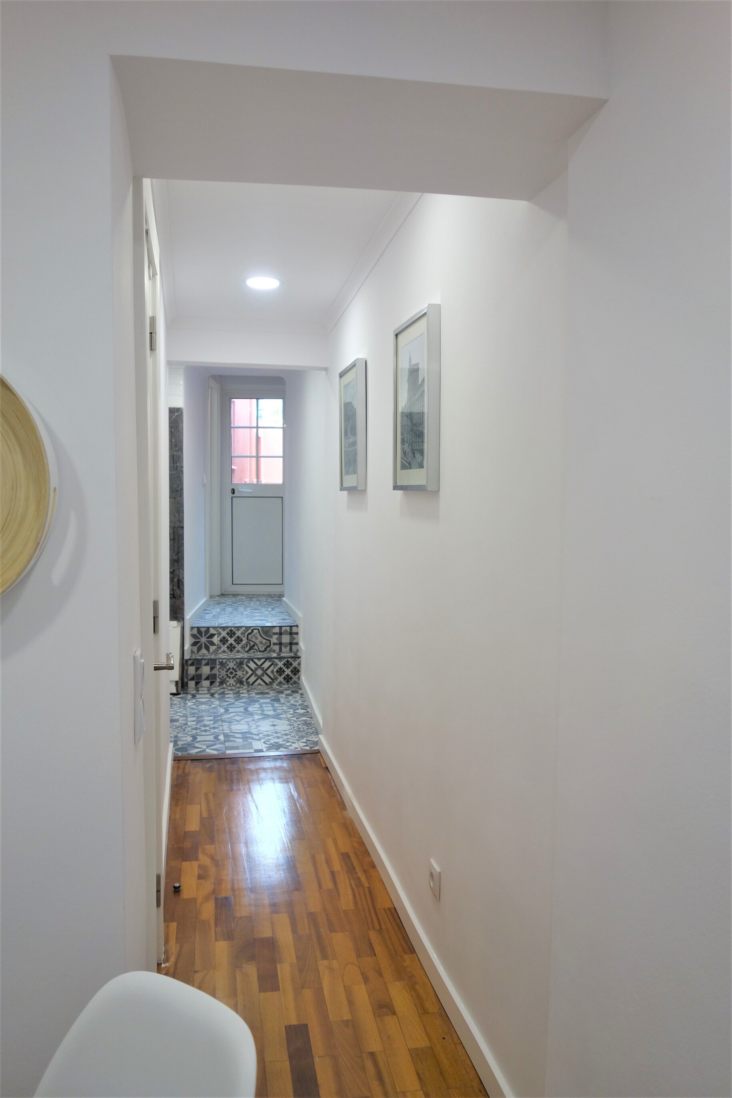 A hallway with a wooden floor, white walls, and a door at the end leading to a room with a patterned tile floor.