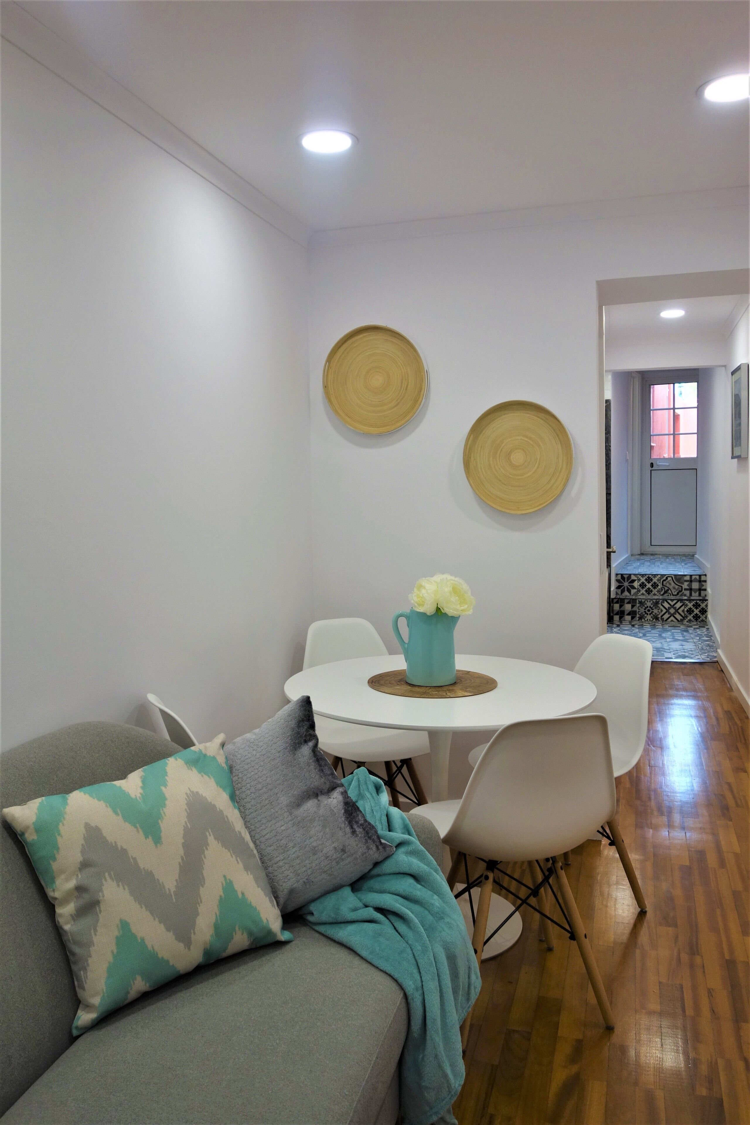 A modern living room with a grey sofa, white chairs, a small round table with a blue pitcher of flowers, and two wall-mounted circular decorations.