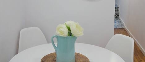 Comedor con una mesa redonda blanca, cuatro sillas blancas, una jarra turquesa con flores blancas y dos adornos tejidos para la pared.