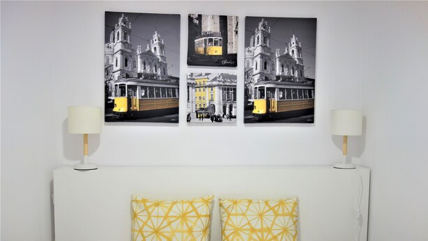 A bed with white headboard and two yellow patterned pillows, a white bedside table with a lamp, and three black and white photographs of a tram and a building on the wall. - Santa Marta Apartment by Trip2Portugal (Lisbon)