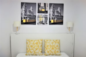 Un lit avec une tête de lit blanche et deux coussins à motifs jaunes, une table de chevet blanche avec une lampe, et trois photographies en noir et blanc représentant un tramway et un bâtiment, accrochées au mur.