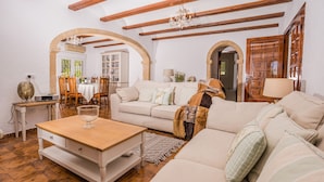 A living room with a wooden coffee table, a beige sofa, and a white armchair. There is a wooden door and a window with a view of greenery. - 3 Bedroom Family Villa in Javea (Javea / Xàbia)