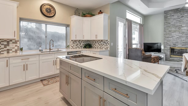 A modern kitchen with white cabinets, a marble countertop, and a built-in oven. There is a large window with blinds, a stone wall, and a television in the living area. - FANTASTIC Scottsdale location! Close to Waste Management Open, and Westworld!! (Scottsdale)