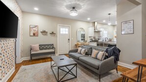 A living room with a sofa, coffee table, and a dining area with a table and chairs. There is a kitchen in the background with cabinets and a countertop. - 3 KING Beds + Walk to River Market + Coffee (Kansas City)
