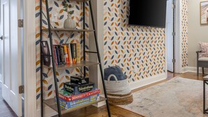 A living room with a bookshelf, a flat-screen TV mounted on the wall, and a patterned wallpaper. - 3 KING Beds + Walk to River Market + Coffee (Kansas City)
