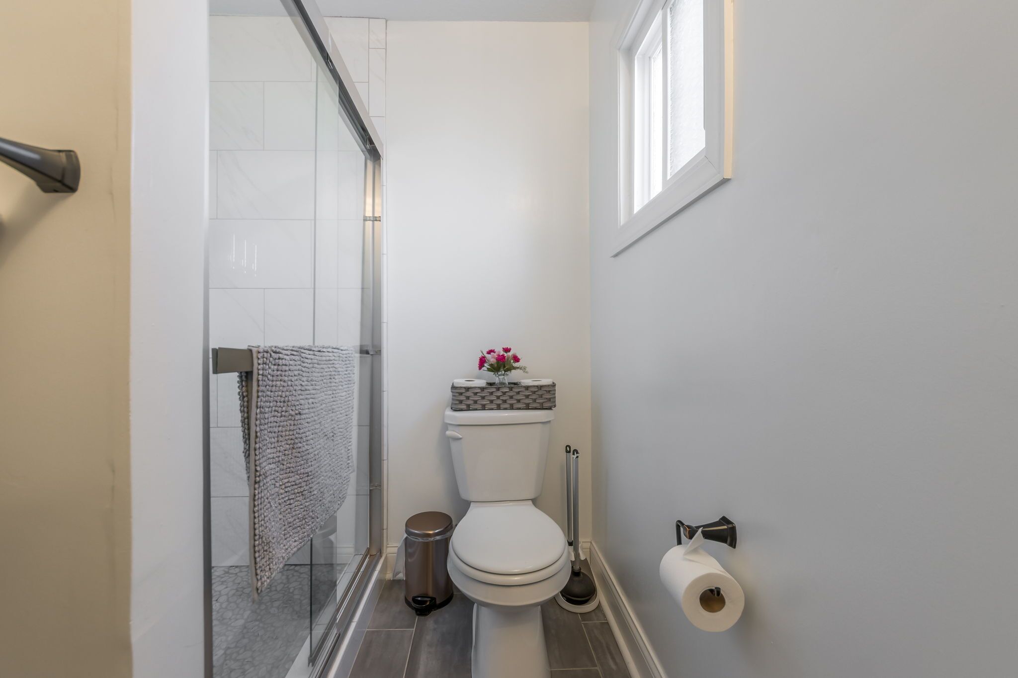 A bathroom with a toilet, a trash bin, a shower, and a toilet paper holder.