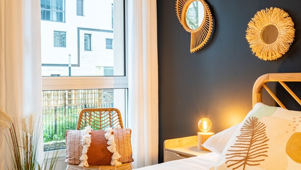 Una camera da letto con un'ampia finestra, un letto con cuscino bianco e decorato, una lampada da comodino e due specchi fissati alla parete.