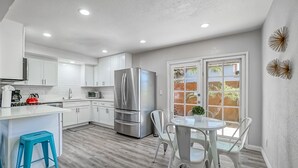 A modern kitchen with white cabinets, a stainless steel refrigerator, and a dining area with white chairs and a table. - Funtierland + Adventure House + Close to Disney + Renovated+ Pool (Anaheim)