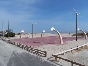 An outdoor basketball court with a red surface, surrounded by sand and wooden railings.