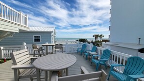 A deck with outdoor seating, a view of the ocean, and a building with a white facade.