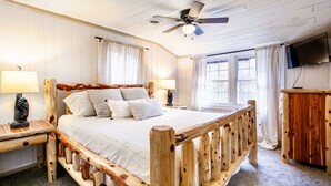 Una cama con estructura de madera, ropa de cama blanca, un ventilador de techo y un televisor en la pared.