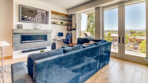A modern living room with a large blue sofa, a flat-screen TV mounted on the wall, a fireplace, and a large sliding glass door leading to a balcony with a city view.