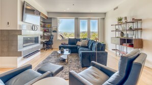 A modern living room with a fireplace, comfortable seating, a bookshelf, and large windows offering a view of the outdoors.