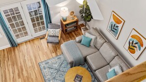 A modern living room with a grey sectional sofa, a round coffee table, and a patterned rug. There is a wooden side table with a lamp and decorative items. A potted plant is on a stand, and artwork is on the wall. - Near downtown Cary & RDU. Easy access to Raleigh, Durham and local venues. (Cary)