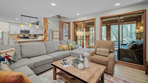 A spacious living area with a grey sectional sofa, a wooden coffee table, and a comfortable armchair. A kitchen with white cabinets and a microwave is visible in the background.