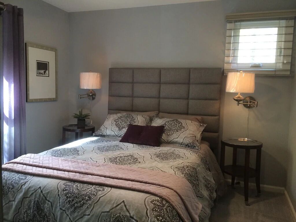 Une chambre à coucher avec un grand lit, deux tables de chevet, une tête de lit et une fenêtre avec des stores.