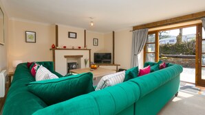 A living room with a green sofa, a fireplace, a TV, and a large window with curtains. - Riverside@the West Dry (St Austell)