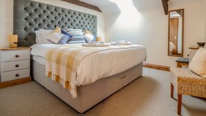 A bedroom with a tufted headboard, a bed with white bedding and multiple pillows, a bedside table with a lamp, a wicker chair, and a skylight. - Water Wheels@the West Dry (St Austell)