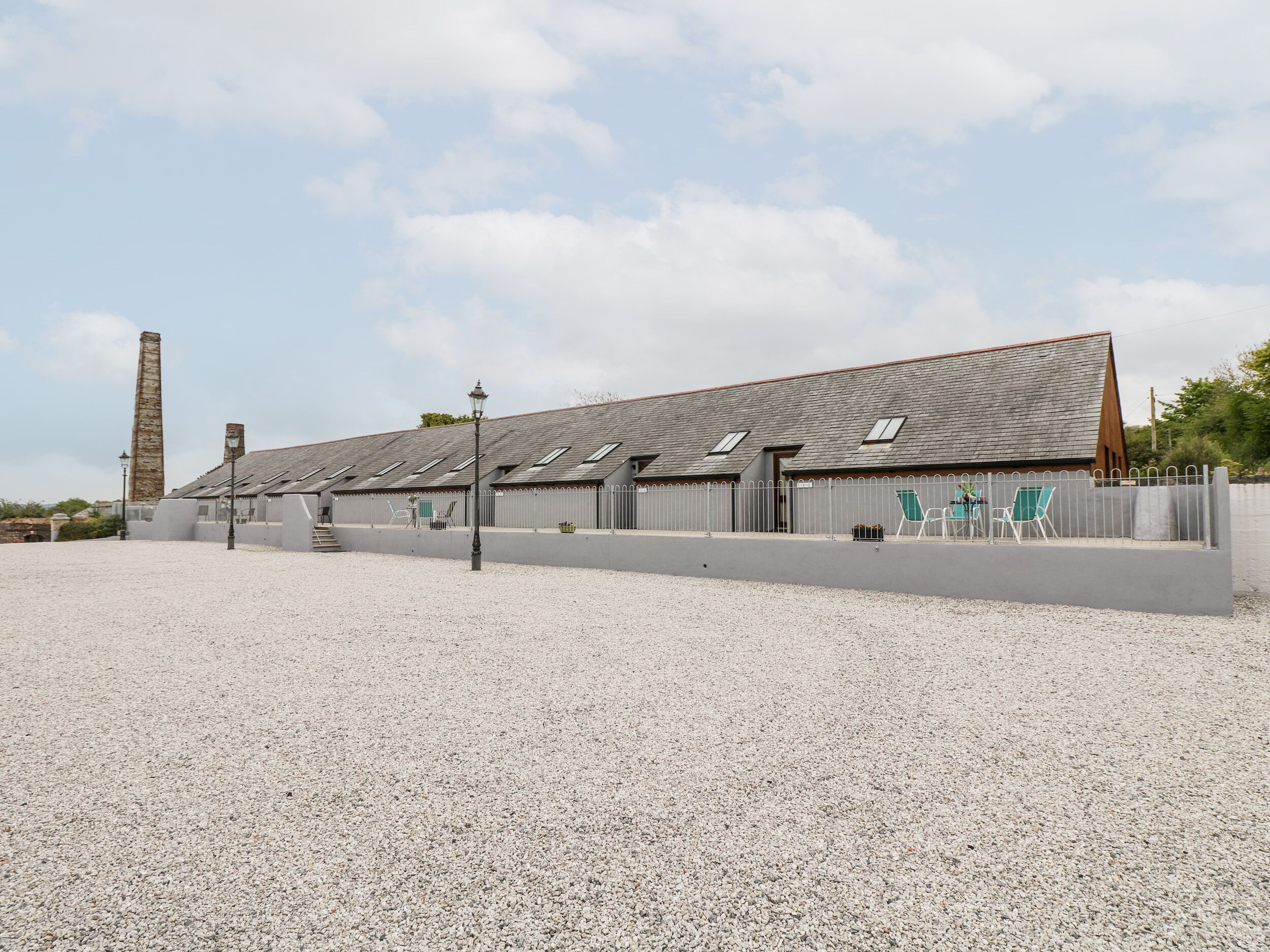 A building with a gravel driveway, a tall chimney, and a fence with chairs and tables.