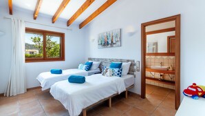 A bedroom with two beds, a window with curtains, a painting on the wall, and a bathroom visible through an open door. - Vista Verde by Balearic Villas (Pollensa)