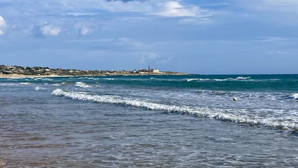 A beach with waves, a clear sky, and a distant shoreline with buildings.