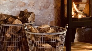 A rustic interior with a stone wall, a fireplace with logs, and a metal basket filled with firewood.