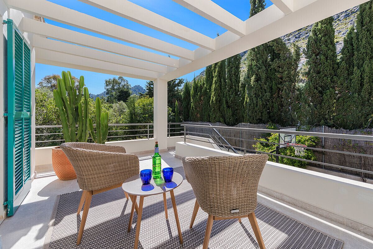 A balcony with wicker chairs and a table, overlooking a mountainous landscape with cacti and trees.