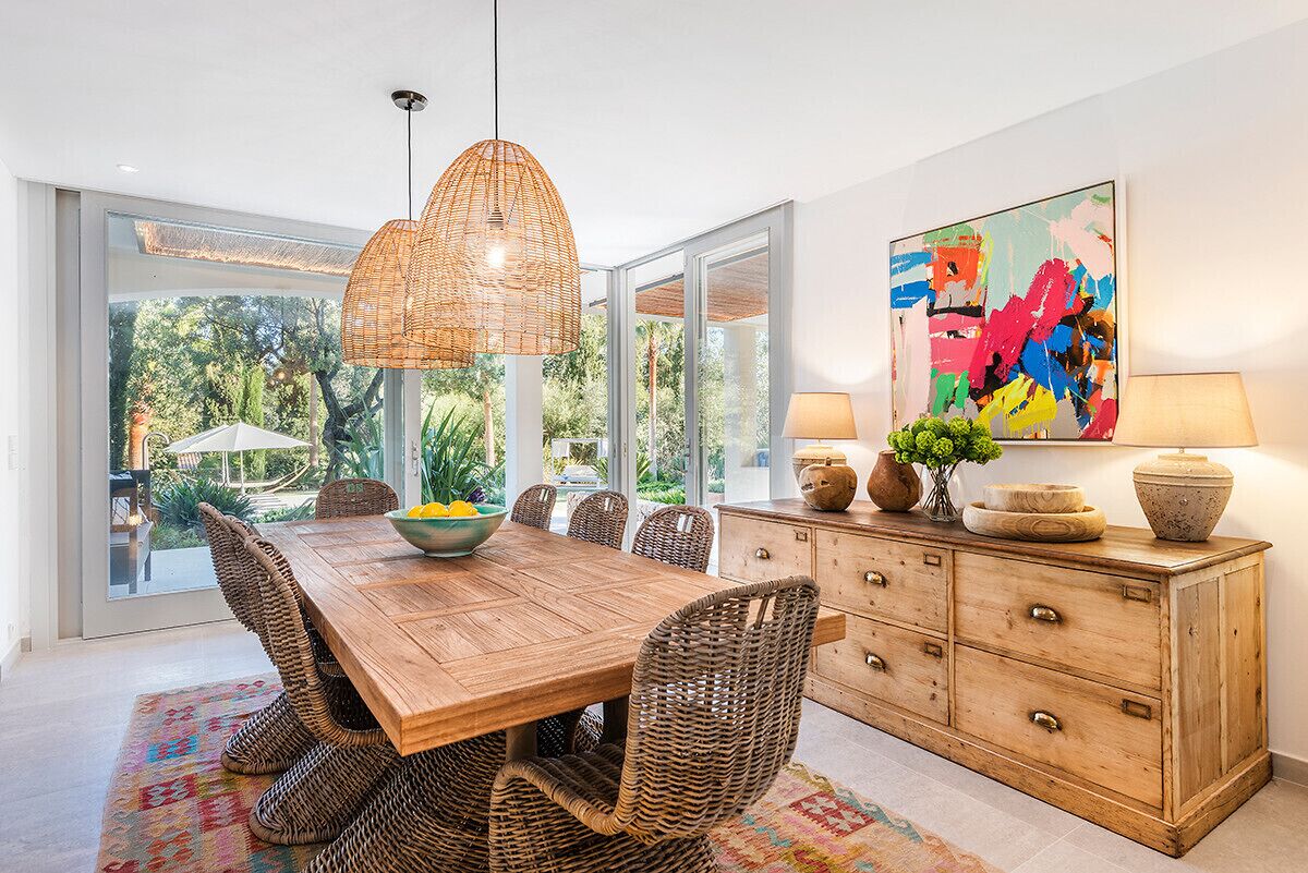 A dining area with a wooden table, wicker chairs, a hanging light fixture, a colorful abstract painting, and a cabinet with decorative items.