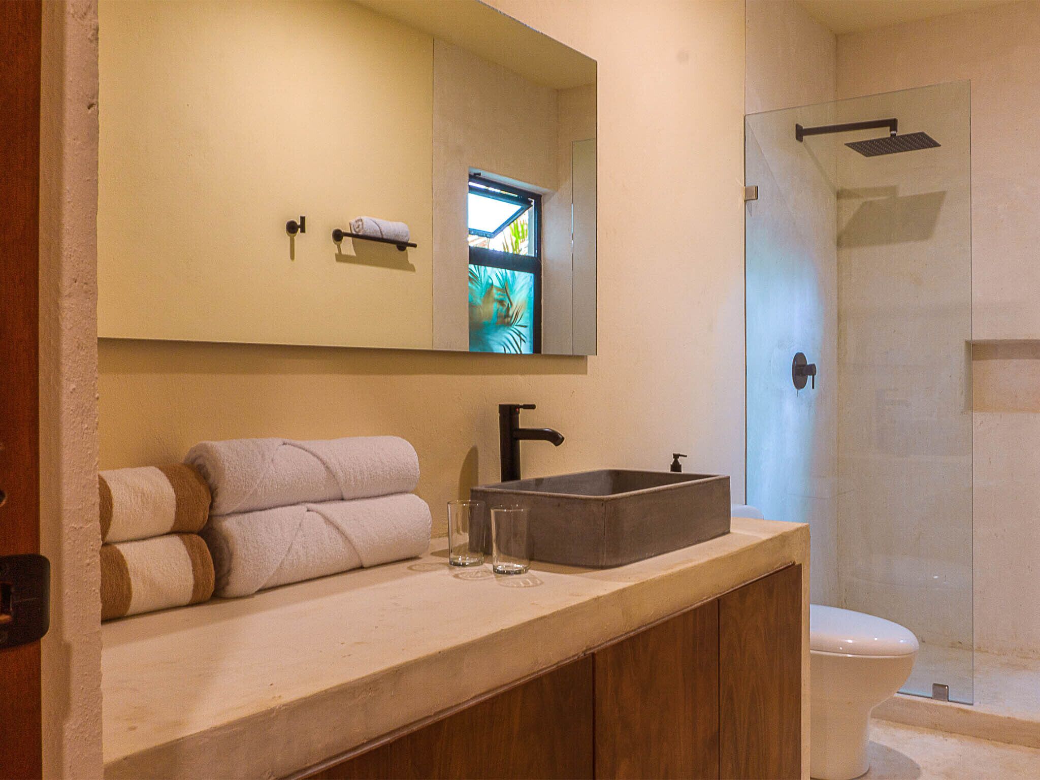 A bathroom with a large mirror, a sink with a rectangular basin, and a toilet. There are towels stacked on the counter and a glass of water.