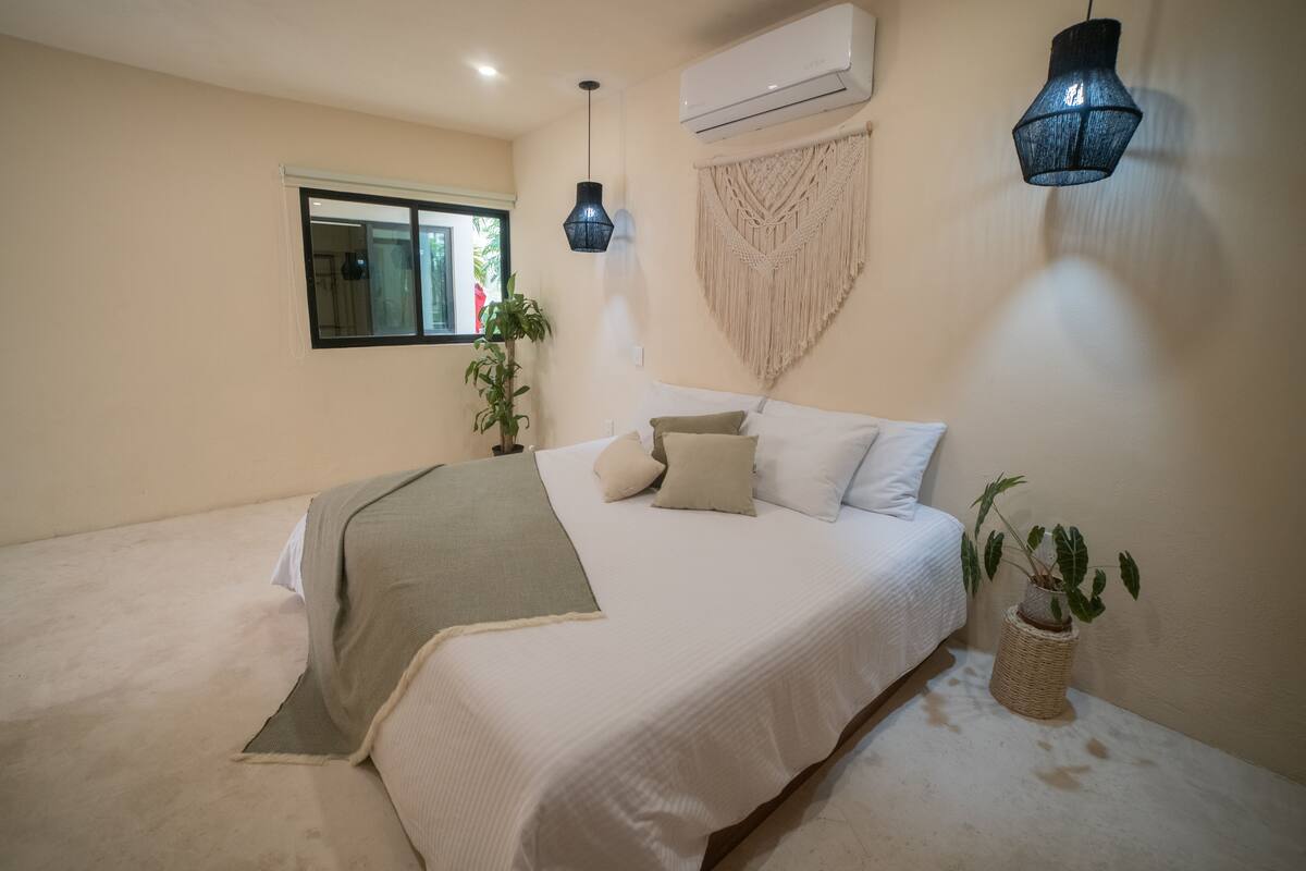 Une chambre à coucher comprenant un lit, une fenêtre, un climatiseur, une décoration murale en macramé et un luminaire suspendu.