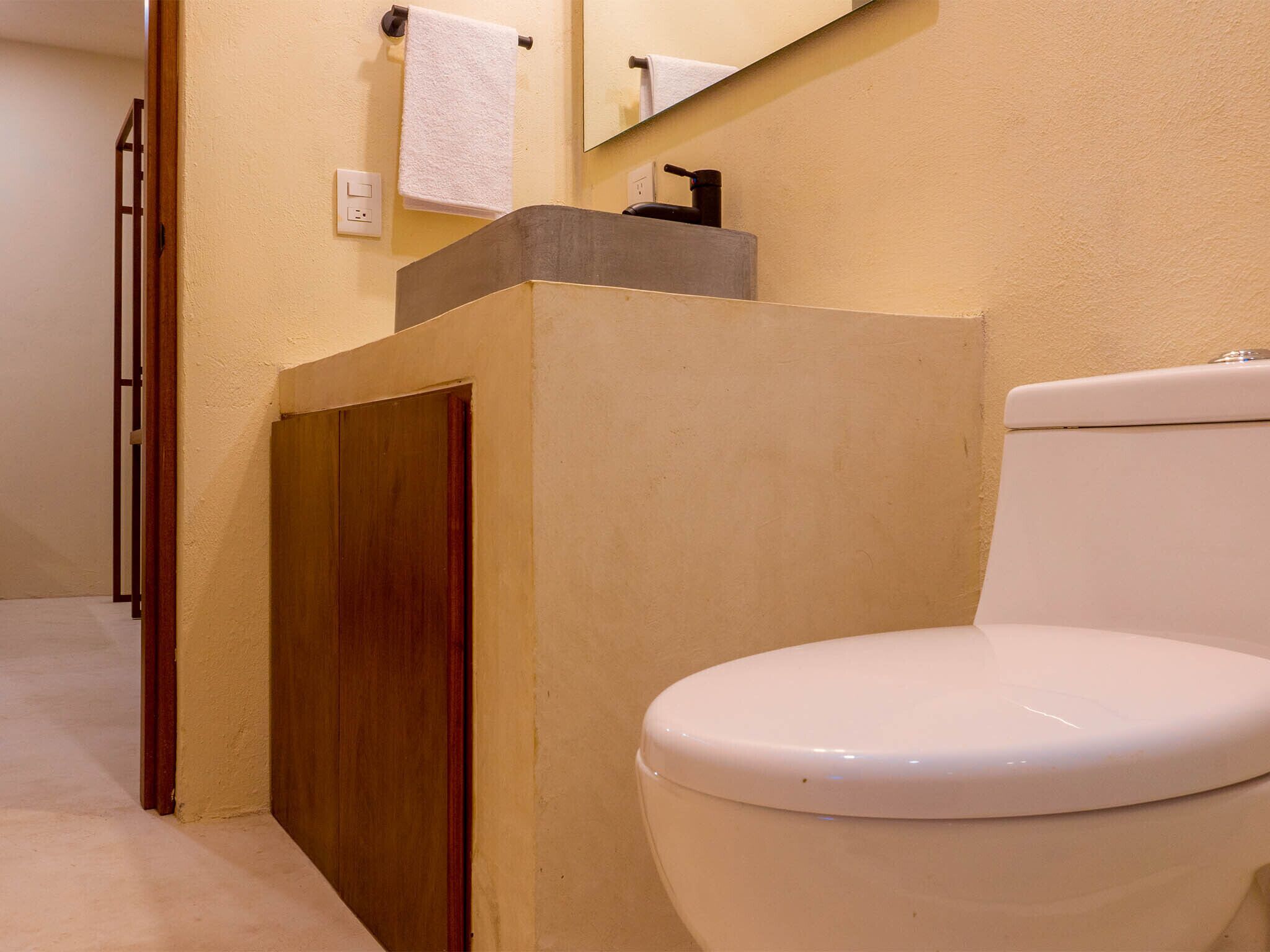 A bathroom with a white toilet, a wooden door, a beige wall, and a mirror above the sink.