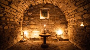 Una stanza in pietra scarsamente illuminata, con un tavolo al centro, due candele e una croce appesa al muro.