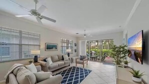 A spacious living room with a large sofa, a smaller chair, and a coffee table. There is a ceiling fan, a TV mounted on the wall, and a large window with blinds. - Beautiful Detached Home in Fiddlers Creek on the Golf Course (Naples)