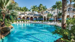 A pool area with lounge chairs and umbrellas, surrounded by palm trees and a building with arched windows. - Beautiful Detached Home in Fiddlers Creek on the Golf Course (Naples)